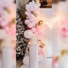 Experience the magic of the heartwarming baby shower decor with a backdrop of pink flex, adorned with a winged neon light and an arch of balloons in white, pastel pink, blush pink, and chrome, creating a whimsical and charming ambiance.