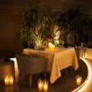 Arrangements for outdoor candlelight dinner with comfortable seating, warm lighting and lush green plants.