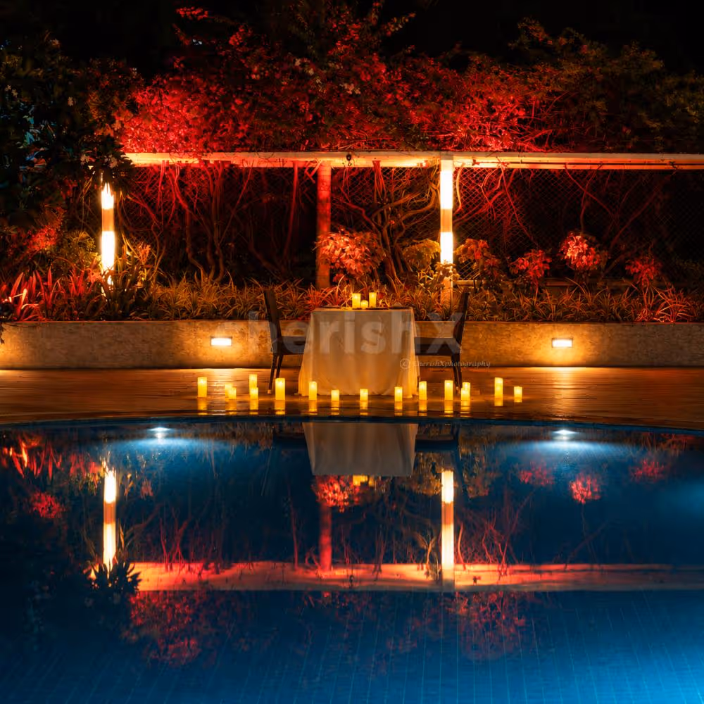 Luxury poolside dining setup with glowing candles, dramatic landscape lighting, and reflections on tranquil blue water.