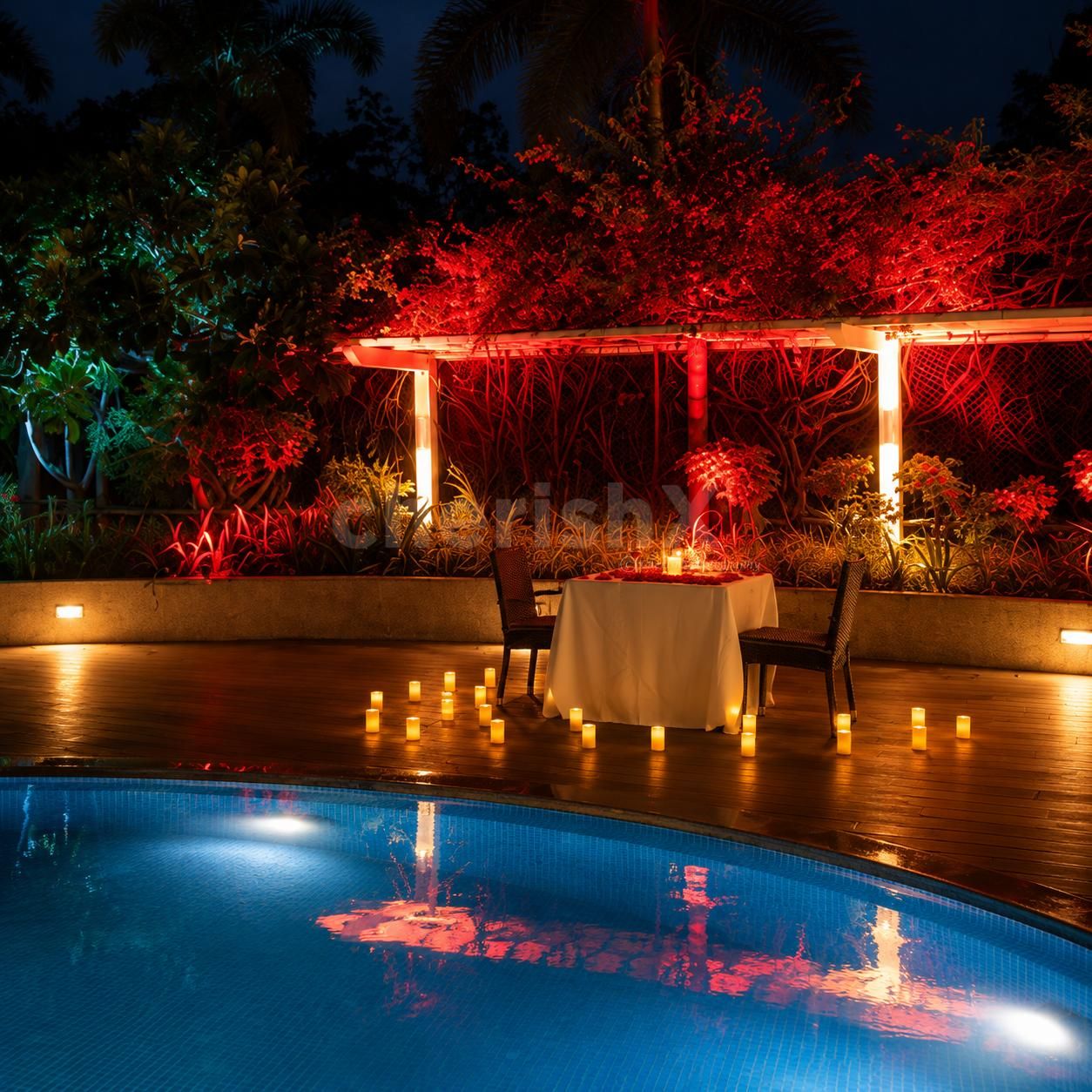 Elegant poolside dinner table decorated with rose petals, wine glasses, glowing candles, and romantic evening lighting.