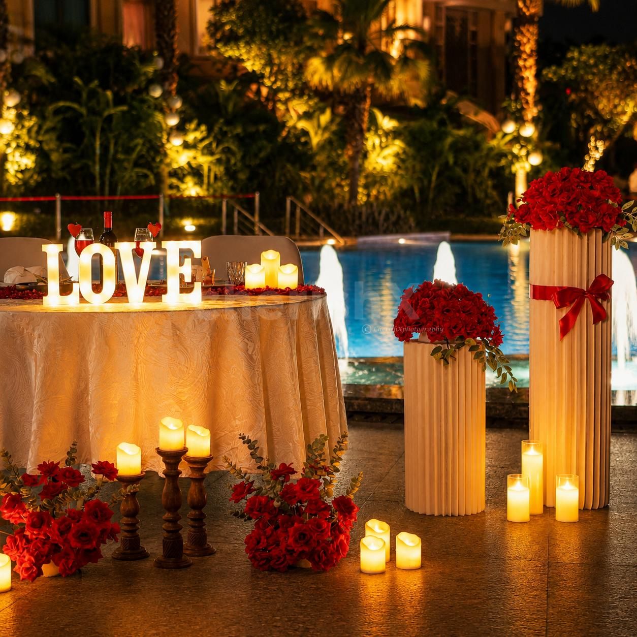 Elegant candlelit poolside dining arrangement featuring red roses, glowing LOVE letters, warm candles, and a beautifully lit blue pool backdrop