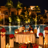 Couple enjoying a romantic poolside dining experience surrounded by red roses, candlelight, and a softly illuminated pool under the night sky