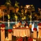 Couple enjoying a romantic poolside dining experience surrounded by red roses, candlelight, and a softly illuminated pool under the night sky