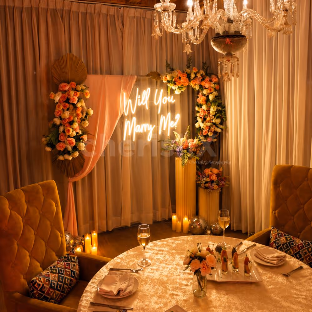 Romantic proposal dinner setup featuring elegant seating, floral arrangements, warm lighting, and a neon "Will You Marry Me?" sign.