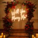 Close Up On Fancy Flower Arrangements Surrounding A Light Up "Will You Marry Me?" Sign.