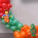The Bottom Part Of The Lego-Themed Decoration Includes Green And Orange Balloon Clusters, Along With Cutouts Of Characters On The Backdrop.
