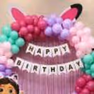 Happy Birthday Gabby's Dollhouse birthday decor with a pink fringe curtain, Happy Birthday bunting, and pastel balloon decor.