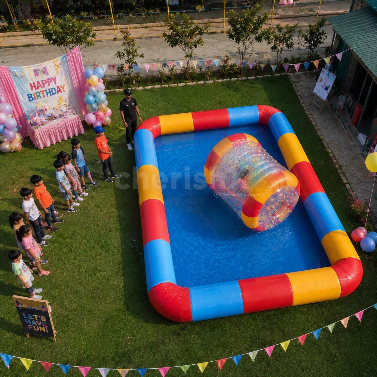 Overhead view of a colorful water zorbing setup at a kids' birthday party with children waiting for their turn and one child enjoying the inflatable water roller.