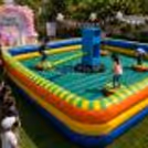 Children spinning arms in a colourful inflatable obstacle eliminator arena at an outdoor birthday party.