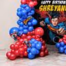 The boundary of the round celebration is dotted with red and blue clusters of balloon bunting and superhero paper cutouts.