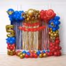 Birthday decoration for Wonder Woman using a red, blue, and golden balloon arch, shimmer foil curtains, themed cutouts, and golden age number balloon.