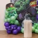 Close-Up Of Hulk Comic Theme Backdrop For Kids Birthday Party With Green And Purple Balloon Garland.