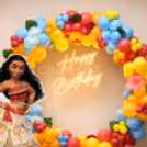 Happy Birthday neon sign with a Moana character standee in a warm white balloon arch inside.