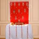 Red velvet puja backdrop, golden lotus hanging, and a white table decorated.