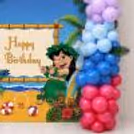Pink and white themed birthday backdrop with pastel balloons and green palm leaves for children's party.
