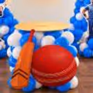 Large red cricket ball & bat cutouts, round table & blue & white balloon clusters for a cricket birthday party.