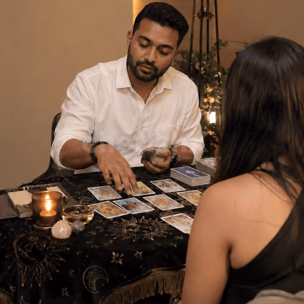 Indian male tarot card reader in a white shirt giving a tarot reading to a female client, with tarot cards, candles and crystals on the table.