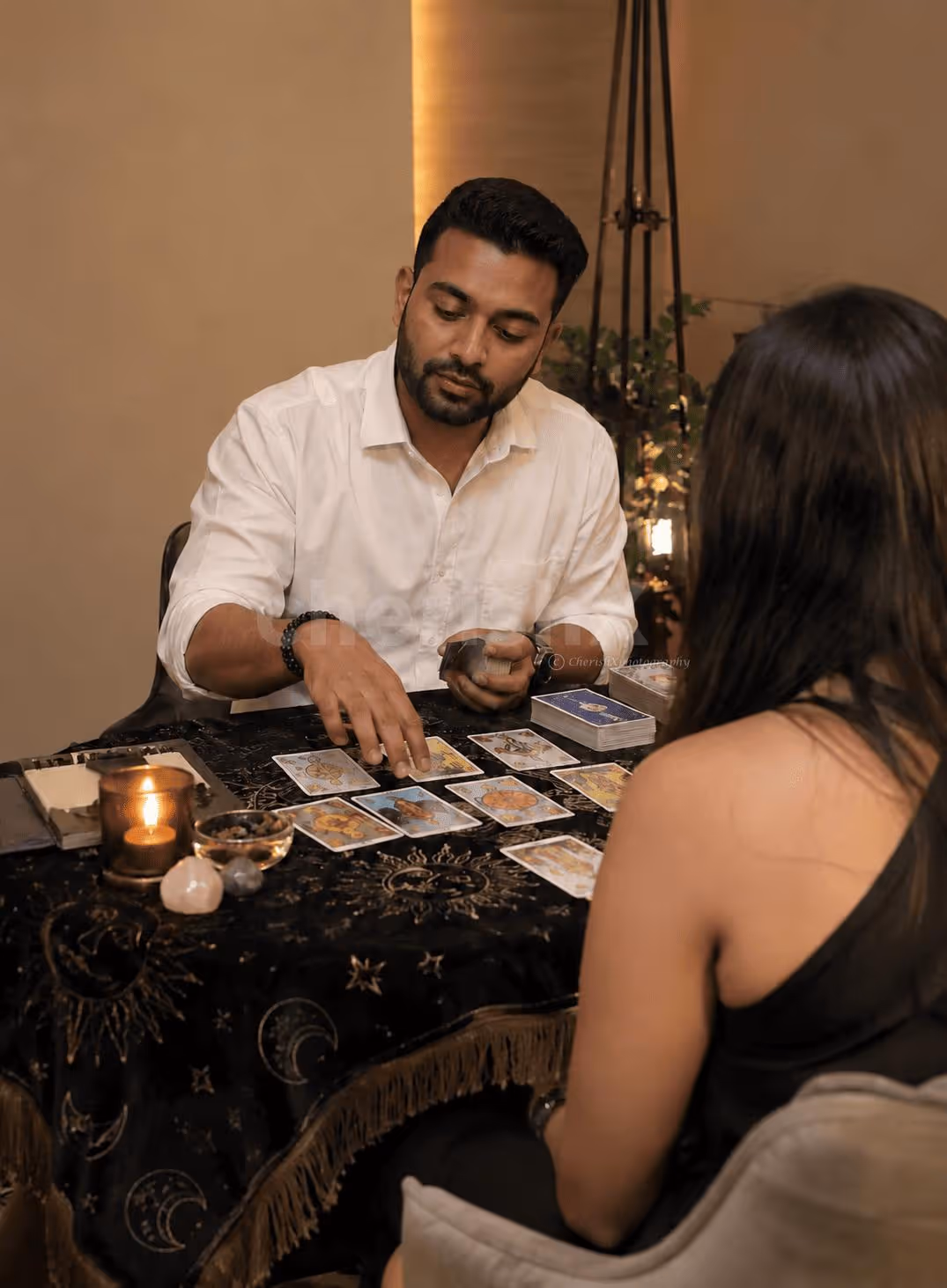 Indian male tarot card reader in a white shirt giving a tarot reading to a female client, with tarot cards, candles and crystals on the table.