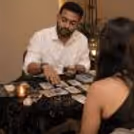 Indian male tarot card reader in a white shirt giving a tarot reading to a female client, with tarot cards, candles and crystals on the table.