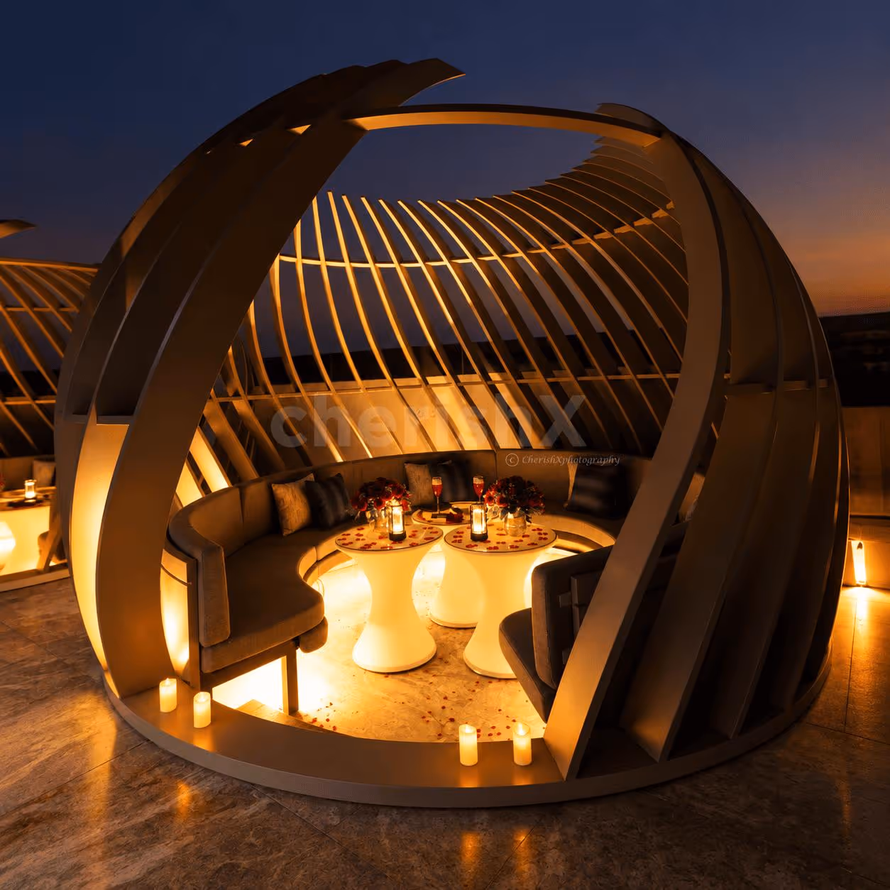 Luxury rooftop gazebo dining area illuminated with warm lights, featuring romantic table settings, flowers and a beautiful night-time city view.