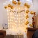 Elegant white and gold balloon birthday decoration with warm fairy lights and a stylish Happy Birthday backdrop, perfect for home birthday celebrations and surprise party setups.
