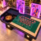 Luxury roulette table setup at an event with a green felt playing surface, roulette wheel, illuminated suit symbols and vibrant purple lighting.