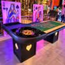 Overhead view of a roulette table with a detailed green game layout, roulette wheel, glowing card-suit decorations and festive event lighting.
