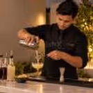 Bartender mixing a drink at a private event bar, with simple bar tools, garnishes and warm ambient lighting in the background.